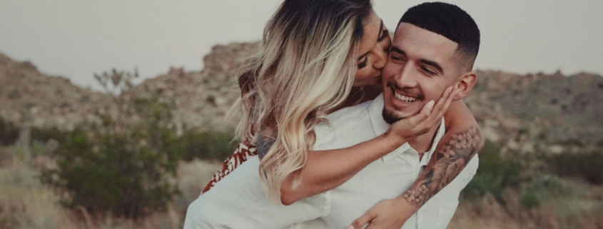 A heterosexual couple in a desert landscape, with the woman riding on the man's back. They're looking at each other affectionately.