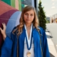 A young white woman in a blue graduation robe and a medal, in front of a painted mural on a building.