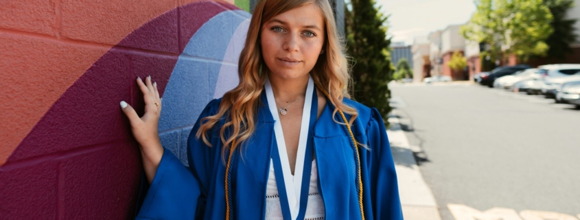 A young white woman in a blue graduation robe and a medal, in front of a painted mural on a building.