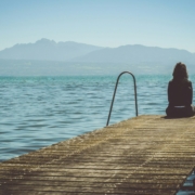 A person sitting at the end of a dock by a lake, with mountains in the background.