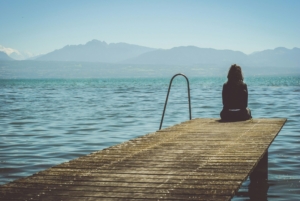 A person sitting at the end of a dock by a lake, with mountains in the background.