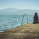 A person sitting at the end of a dock by a lake, with mountains in the background.