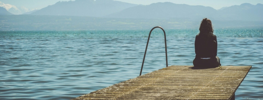 A person sitting at the end of a dock by a lake, with mountains in the background.