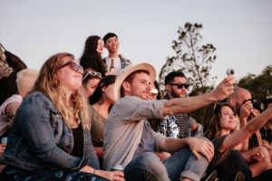 A group of people sitting together outside, while one man in the center holds out his phone for a selfie.