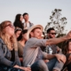 A group of people sitting together outside, while one man in the center holds out his phone for a selfie.