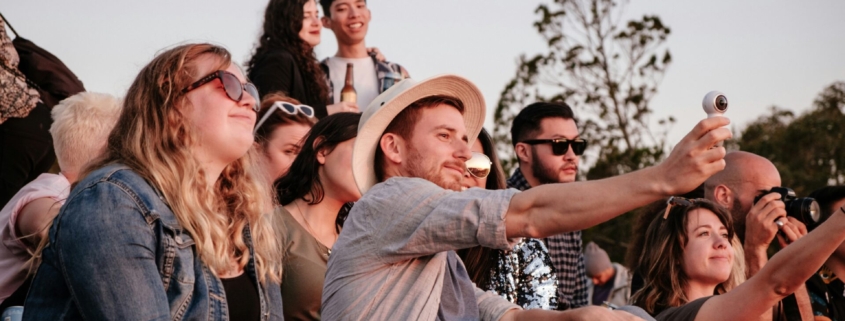 A group of people sitting together outside, while one man in the center holds out his phone for a selfie.