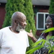 A stock photo of an older Black man talking to a younger Black woman, who is smiling, outside.