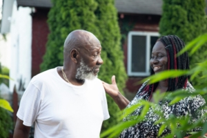 A stock photo of an older Black man talking to a younger Black woman, who is smiling, outside.