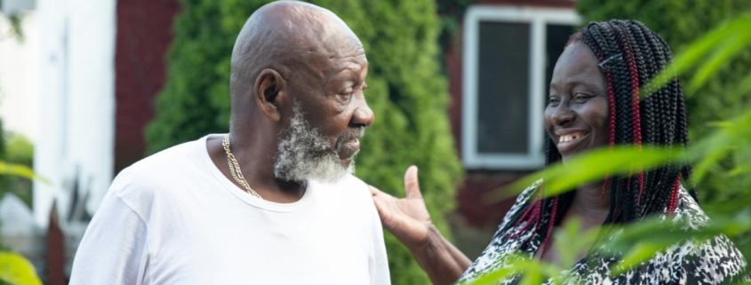 A stock photo of an older Black man talking to a younger Black woman, who is smiling, outside.