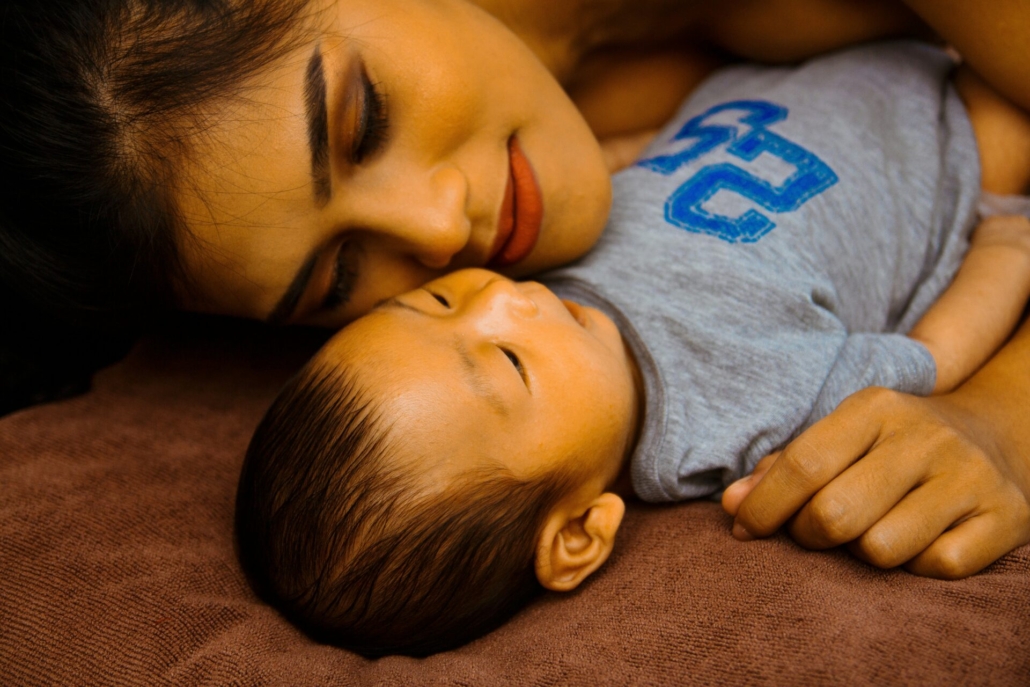 A woman with olive skin and dark brown hair laying on her side, next to an infant with hair the same color as hers.