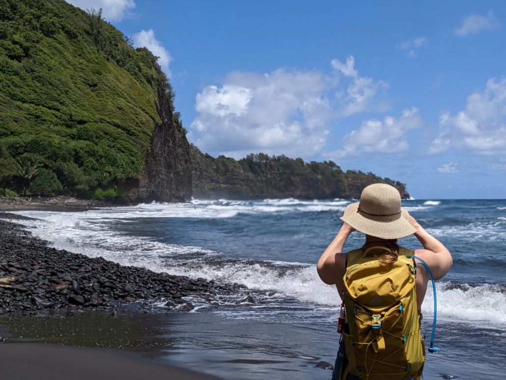 A person wearing a travel backpack and a khaki hat standing on a beach in front of lush cliffs, taking a photo.