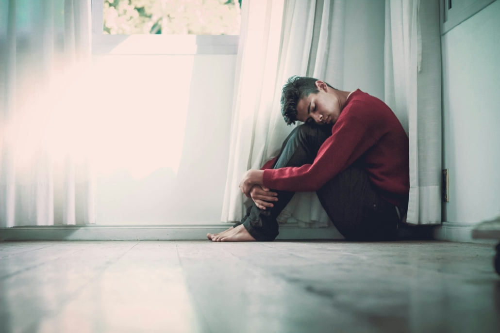 A man sitting on the floor of a gray room, hugging his knees to his chest.