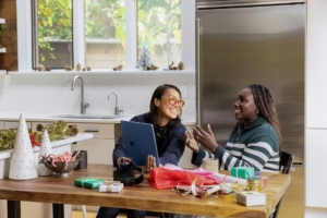 Two women, one of Asian heritage and one Black, sitting together in a kitchen, talking and smiling.