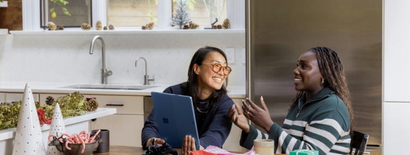 Two women, one of Asian heritage and one Black, sitting together in a kitchen, talking and smiling.