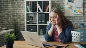 A white woman sitting in front of a laptop at a table, with her head in her hands looking stressed.