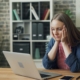 A white woman sitting in front of a laptop at a table, with her head in her hands looking stressed.