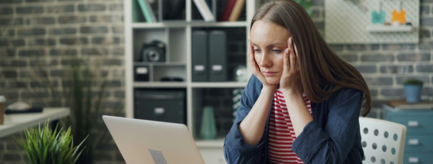 A white woman sitting in front of a laptop at a table, with her head in her hands looking stressed.