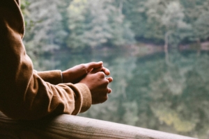 A person's hands and arms, leaning over a railing with water and trees beyond.