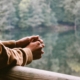 A person's hands and arms, leaning over a railing with water and trees beyond.