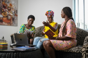 A group of Black women sitting on a couch, talking. They are holding notebooks and there is a computer on the table in front of them.