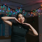 A woman with brown skin and hair, posing in front of a string of Christmas lights.