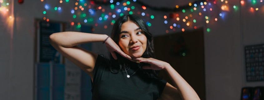 A woman with brown skin and hair, posing in front of a string of Christmas lights.