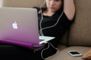 A young white girl using a laptop with corded headphones, with a phone on the couch beside her.