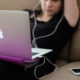 A young white girl using a laptop with corded headphones, with a phone on the couch beside her.