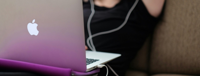 A young white girl using a laptop with corded headphones, with a phone on the couch beside her.