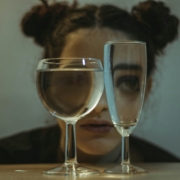 A woman with her hair in space buns crouching behind two glasses filled with water, so her eyes look distorted through the glass.