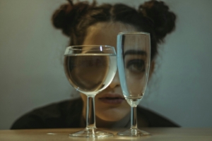 A woman with her hair in space buns crouching behind two glasses filled with water, so her eyes look distorted through the glass.