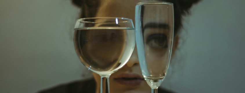 A woman with her hair in space buns crouching behind two glasses filled with water, so her eyes look distorted through the glass.