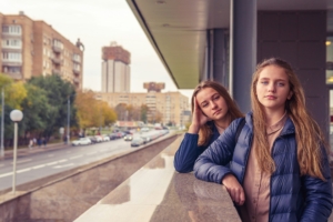 Two bored looking young girls leaning on a ledge, with a city scene in the background.