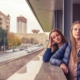 Two bored looking young girls leaning on a ledge, with a city scene in the background.