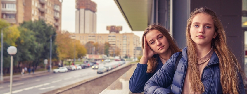 Two bored looking young girls leaning on a ledge, with a city scene in the background.