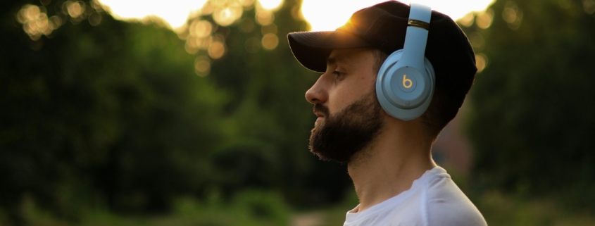 A bearded white man man wearing over the ear headphones outdoors.
