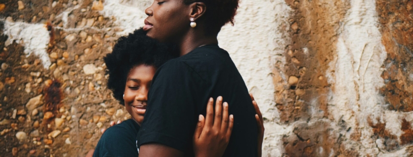 A Black mother hugging her Black teenage daughter.