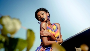 A Black woman in a brightly colored dress hugging herself against a blue sky, with some plants in the foreground.