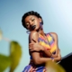 A Black woman in a brightly colored dress hugging herself against a blue sky, with some plants in the foreground.