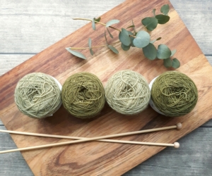 4 balls of green yarn, a pair of knitting needles, and a sprig of eucalyptus on a wooden cutting board.