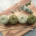 4 balls of green yarn, a pair of knitting needles, and a sprig of eucalyptus on a wooden cutting board.