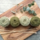4 balls of green yarn, a pair of knitting needles, and a sprig of eucalyptus on a wooden cutting board.