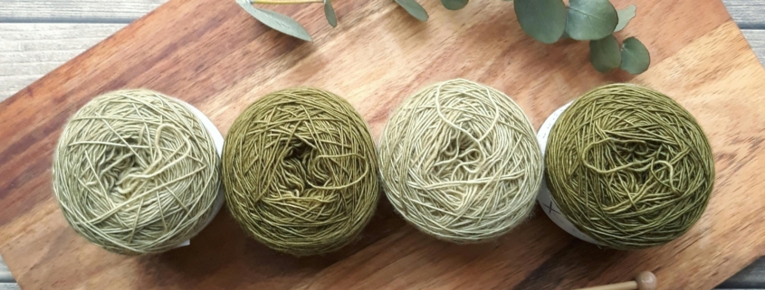 4 balls of green yarn, a pair of knitting needles, and a sprig of eucalyptus on a wooden cutting board.