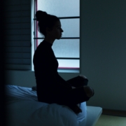 A silhouette of a woman sitting on the end of a bed in a dark room.