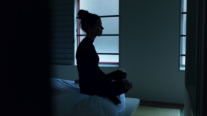 A silhouette of a woman sitting on the end of a bed in a dark room.