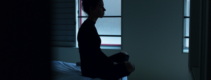 A silhouette of a woman sitting on the end of a bed in a dark room.