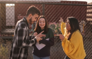 A group of young adults standing outside, looking at a phone and talking.