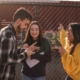 A group of young adults standing outside, looking at a phone and talking.