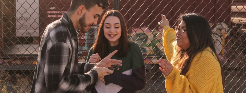 A group of young adults standing outside, looking at a phone and talking.