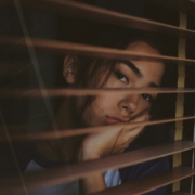 A sad looking young woman looking out a window through the open blinds.